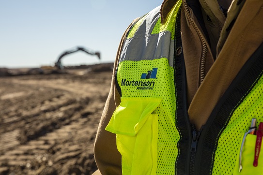 Photo of Mortenson Team member wearing yellow vest and excavator in background.